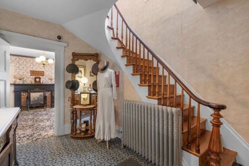 A vintage hallway with a staircase, a radiator, and a mirror displaying hats, leading to a cozy living area.