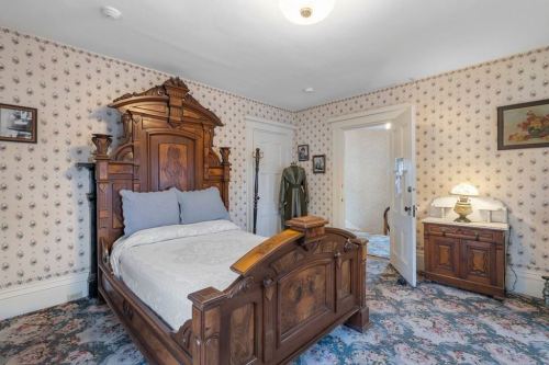 A vintage bedroom featuring an ornate wooden bed, floral wallpaper, and a small nightstand with a lamp.