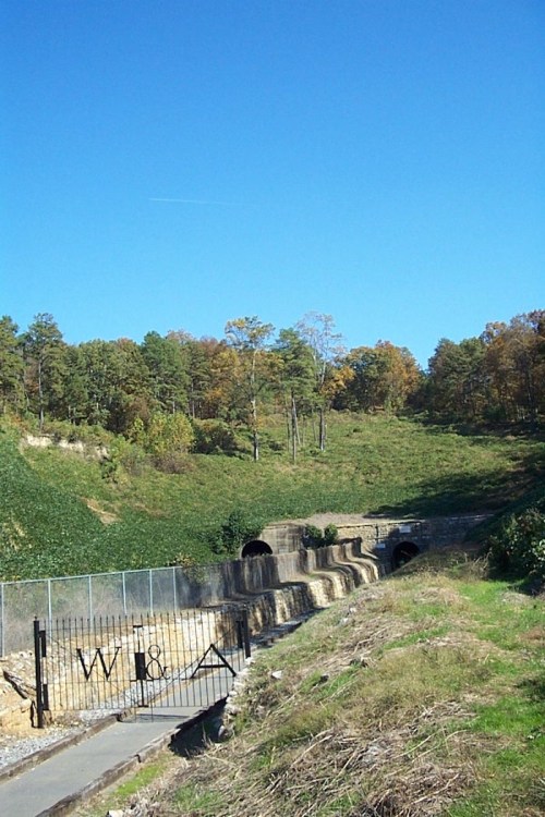 A landscape featuring a grassy hill with trees, two tunnel entrances, and a gated pathway under a clear blue sky.