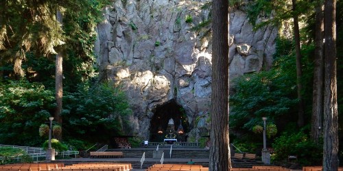 A natural rock formation with a cave entrance, surrounded by tall trees and an empty outdoor amphitheater.
