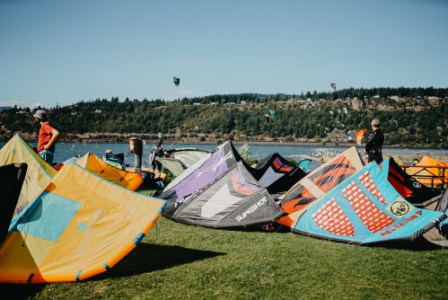 Colorful kitesurfing sails on grass by a lake, with people preparing for kitesurfing in the background.