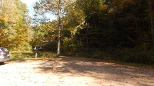 A gravel parking area surrounded by trees with autumn foliage, featuring two people walking in the distance.