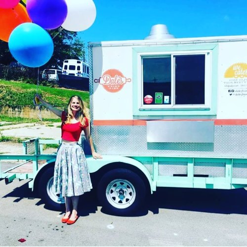 A woman in a red top and floral skirt stands by a food truck, holding colorful balloons against a clear blue sky.