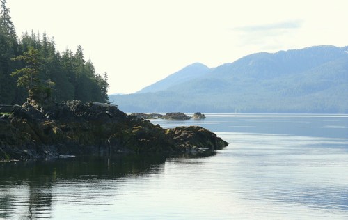 Serene coastal landscape with rocky shores, calm waters, and distant mountains under a clear sky.