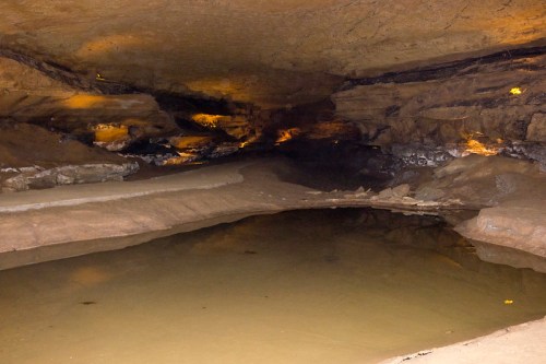 A dimly lit cave interior with a calm water pool reflecting the rocky surroundings.