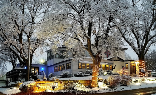 A cozy farmhouse restaurant illuminated by lights, surrounded by snow-covered trees on a winter evening.