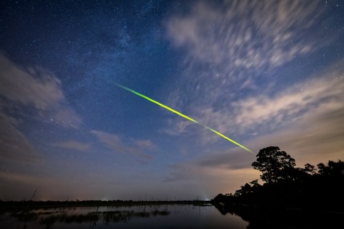 A vibrant green meteor streaks across a starry night sky over a calm lake, surrounded by trees and clouds.