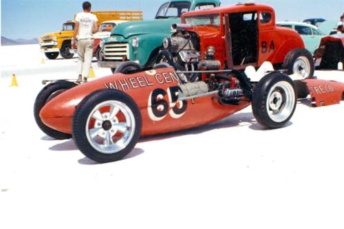 A vintage race car with a red body and large wheels, parked on a salt flat with other classic vehicles in the background.