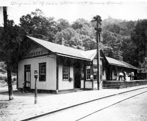 Historic black-and-white photo of a train station with a wooden structure and people on the platform, surrounded by trees.