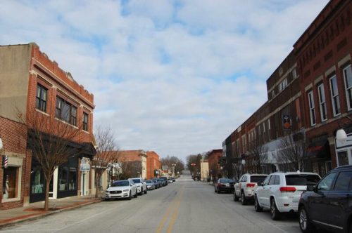 A quiet downtown street lined with brick buildings and parked cars under a cloudy sky.