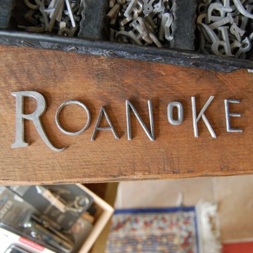 Metal letters spelling "ROANOKE" on a wooden surface, with various other letters in the background.