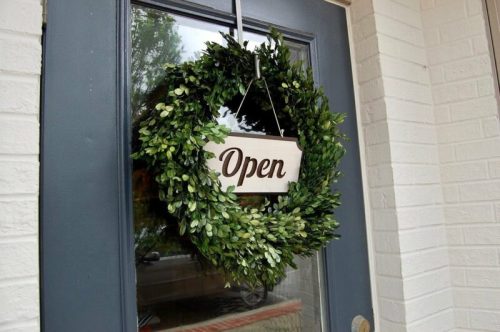 A green wreath with an "Open" sign hangs on a blue door, welcoming visitors.