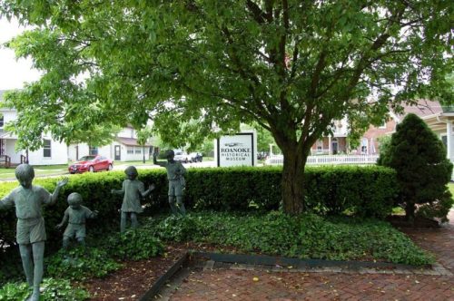A green park area with a tree, a sign for Roanoke Historical Museum, and bronze child sculptures playing.