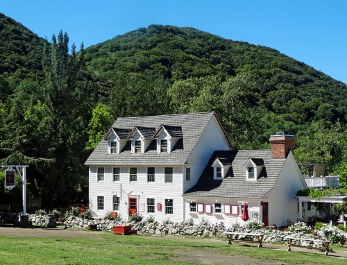 A charming white house with a steep roof, surrounded by greenery and mountains, featuring a stone wall and flower beds.