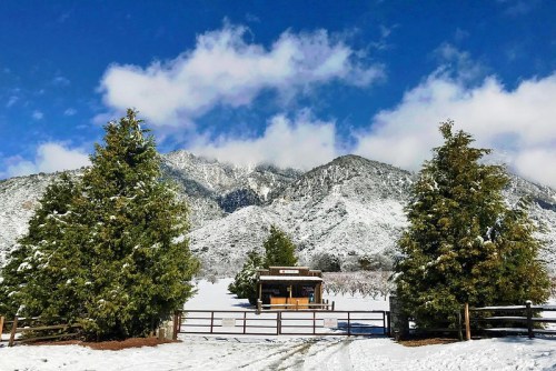 A snowy landscape with evergreen trees, a small cabin, and mountains under a blue sky with clouds.