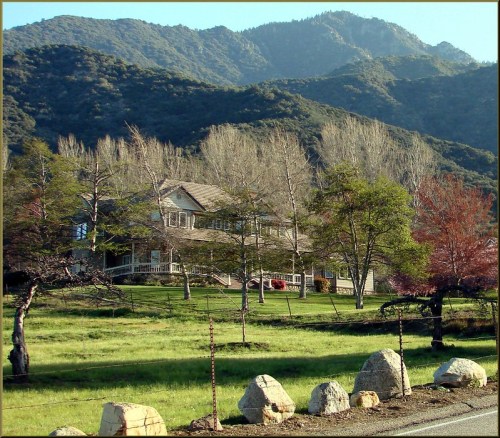 A scenic view of a house surrounded by trees and mountains, with a grassy area and rocky foreground.