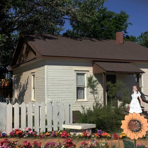 A quaint house with a white picket fence, surrounded by colorful flowers and a cutout of a girl in a white dress.