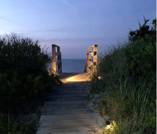 A wooden walkway leads to the beach at dusk, surrounded by grass and bushes, with soft lighting illuminating the path.