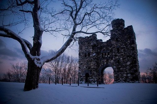 Ruins of a stone castle surrounded by snow, with a bare tree in the foreground and a twilight sky in the background.