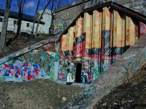 Colorful graffiti covers a concrete structure, with a partially visible entrance and surrounding trees under a blue sky.