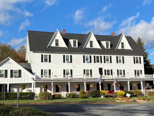 A large white historic building with multiple gables, surrounded by greenery and colorful flower pots under a blue sky.