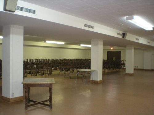 A large, empty room with stacked chairs on tables and a few tables scattered around, illuminated by overhead lights.