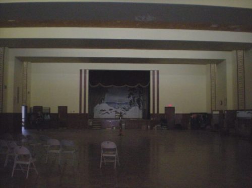 Empty hall with a stage at the front, featuring a painted backdrop and rows of folding chairs set up.