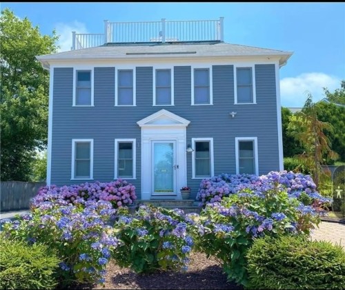 A blue two-story house with a white door, surrounded by colorful hydrangea flowers in bloom.