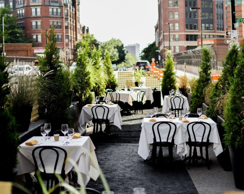 Outdoor dining area with neatly set tables and chairs, surrounded by greenery and city buildings in the background.