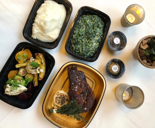 A table setting with containers of mashed potatoes, sautéed greens, roasted vegetables, and a grilled steak, plus candles.