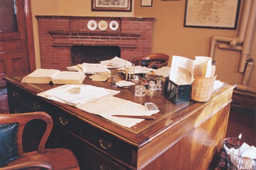A cluttered wooden desk with papers, ink bottles, and a basket, set in a cozy room with a brick fireplace.