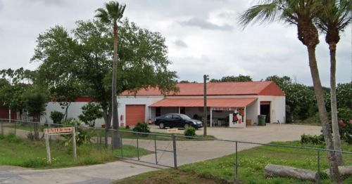 A single-story building with a red roof, surrounded by palm trees and a fence, featuring a parking area and entrance sign.