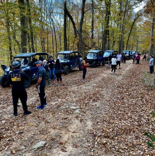 A group of people stands along a dirt path with off-road vehicles in a wooded area during autumn.