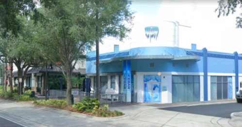 A blue and white building with a sign, surrounded by trees and a sidewalk, in a sunny urban setting.
