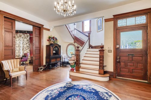 Elegant foyer with a staircase, wooden accents, a chandelier, and a decorative rug. Natural light streams in through windows.