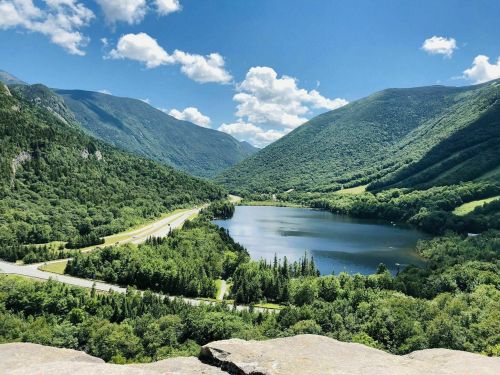 A scenic view of a lake surrounded by lush green mountains under a bright blue sky with fluffy clouds.