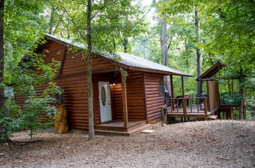 A rustic wooden cabin surrounded by trees, featuring a porch and a small deck.