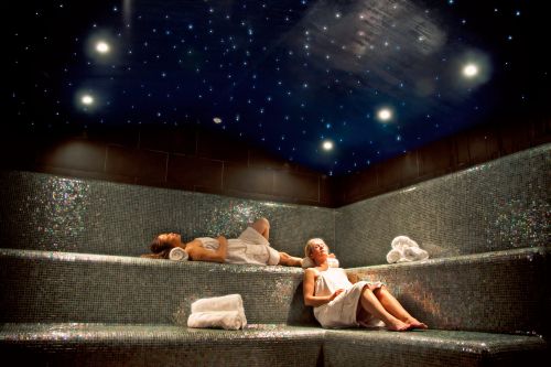 Two people relax in a serene spa setting with starry ceiling and mosaic-tiled benches, surrounded by rolled towels.