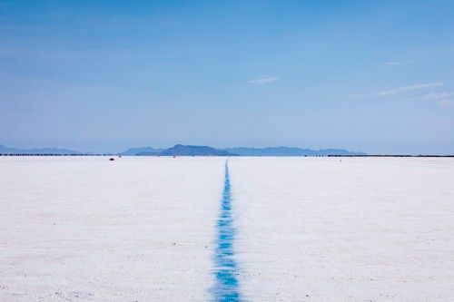 A vast white salt flat stretches into the distance, with a faint blue line leading towards distant mountains under a clear sky.