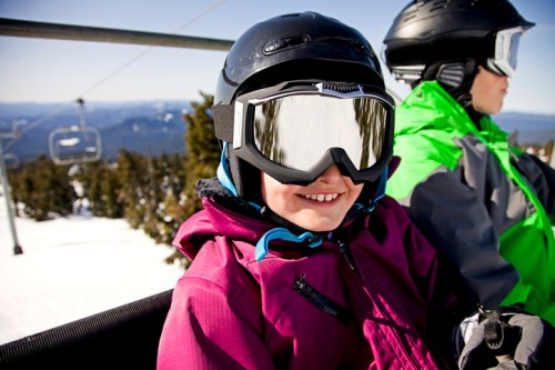 A smiling child in a purple jacket and helmet rides a ski lift, with snowy mountains in the background.