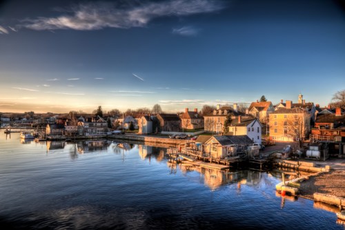 A serene waterfront scene at sunset, featuring charming houses and boats reflecting in calm water.