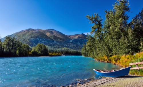 A serene river scene with a blue boat, surrounded by lush trees and mountains under a clear blue sky.