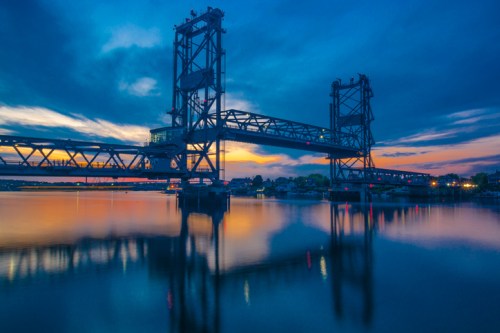 A bridge over calm water at sunset, reflecting vibrant colors in the sky and water below.
