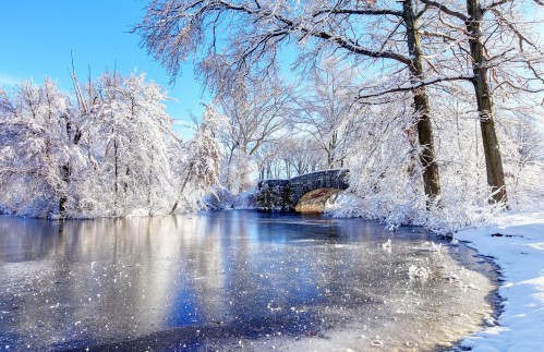 A serene winter scene featuring a frozen river, snow-covered trees, and a stone bridge under a clear blue sky.