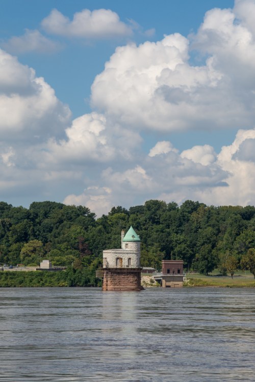 A small stone tower with a green dome stands in a river, surrounded by lush trees and a blue sky with fluffy clouds.