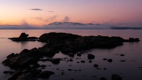 A serene landscape at dawn, featuring rocky shores and snow-capped mountains shrouded in mist under a pastel sky.