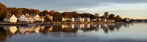 A serene waterfront scene with charming houses reflecting in calm water during sunset. Trees in autumn colors line the shore.