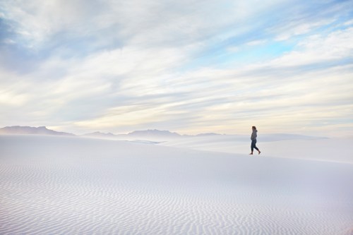 A person walks alone across a vast, white sand dune landscape under a soft, cloudy sky.