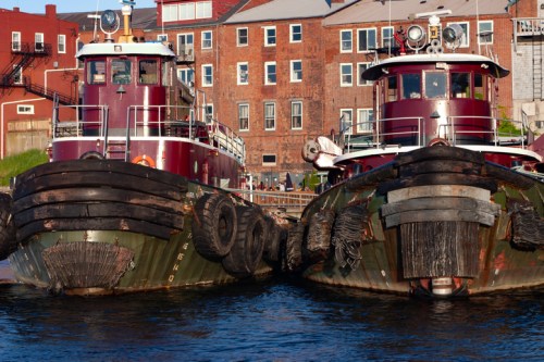 Two tugboats moored side by side in a harbor, with brick buildings in the background and clear blue water.