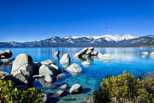 A serene lake scene with clear blue water, rocky formations, and snow-capped mountains under a bright blue sky.